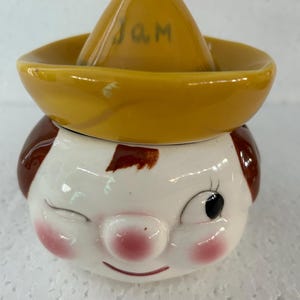 Peut inclure: Un pot à confiture en céramique en forme de visage de dessin animé avec un chapeau brun. Le chapeau porte l'inscription "JAM". Le visage a un œil qui cligne, un nez rose et des joues roses.