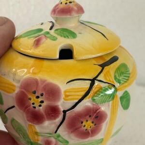 Peut inclure: Un petit sucrier rond en céramique avec un couvercle. Le bol est peint avec des rayures jaunes et blanches, et présente des fleurs roses et des feuilles vertes. Le couvercle a un petit bouton et une fente pour une cuillère.