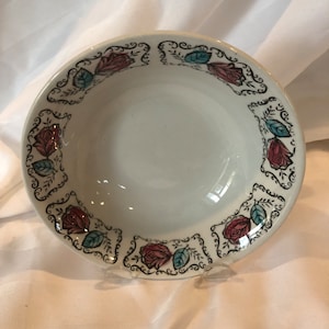 May include: A white ceramic bowl with a floral design. The rim features a repeating pattern of red roses, teal leaves, and black scrollwork. The bowl is displayed on a clear stand against a white background.