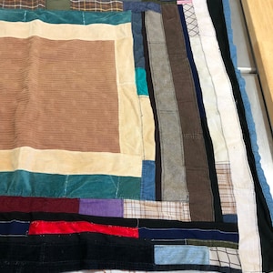 May include: A patchwork quilt made from various fabrics in shades of brown, blue, green, red, and purple. The quilt is made up of squares and rectangles of different sizes and colors, creating a geometric pattern.