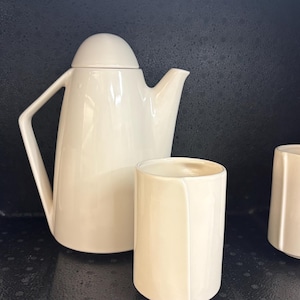 May include: A cream-coloured ceramic tea set, comprising a teapot with a lid, two handled mugs, and two cylindrical cups. The set is arranged on a dark surface, showcasing a minimalist design.