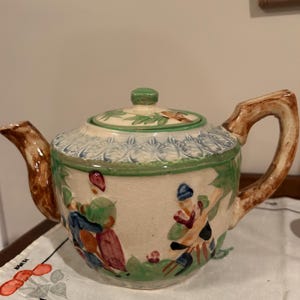 May include: A vintage ceramic teapot with a lid. The cream-coloured body features painted figures and green accents. The handle and spout are brown. The lid has a green knob and floral designs.