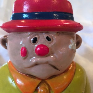 May include: A ceramic clown figurine with a red hat, blue band, and a yellow shirt with an orange collar. The clown has a red nose, red cheeks, and a sad expression. The figurine is painted with glossy paint.