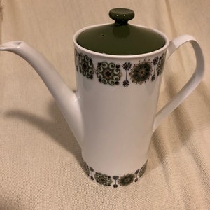May include: White ceramic coffee pot with a green lid and handle. The pot has a decorative band of green and brown floral patterns around the top and bottom. The spout is long and curved.