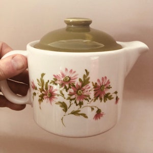 Pode incluir: Cremeira de cerâmica branca com um desenho floral de flores rosa e folhas verdes. A cremeira tem uma tampa verde-oliva e uma pega. É projetada para servir creme ou leite.