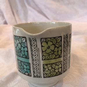 May include: Cream-coloured ceramic creamer with a floral pattern in teal and green. The design is framed by black lines and geometric shapes. The creamer has a small spout and a rounded base.