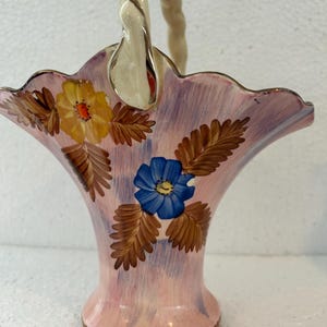 May include: A vintage ceramic basket with a scalloped rim, painted in shades of pink and lavender. The basket features hand-painted yellow and blue flowers with brown leaves. The handle is a twisted cream color with gold accents.