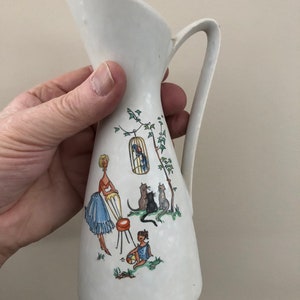 May include: White ceramic pitcher with a handle, featuring a colorful illustration of a woman, a child, and cats. The illustration depicts a whimsical scene with a birdcage and a tree.