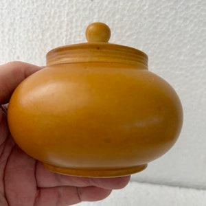 May include: A small, round, yellow-orange jar with a matching lid and a small knob on top. The jar has a smooth, matte finish and a slightly bulbous shape. The lid fits snugly on top, creating a tight seal. The jar is held in a hand.
