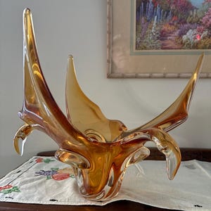 Puede incluir: Un cuenco decorativo de vidrio color ámbar con un diseño abstracto. El cuenco tiene múltiples extensiones puntiagudas y una forma acampanada. El vidrio es transparente, permitiendo el paso de la luz. Está sobre una superficie de madera.