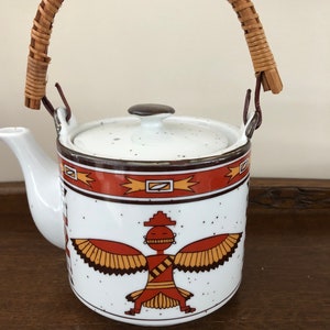 May include: White ceramic teapot with a brown and orange Native American design. The teapot has a woven rattan handle and a matching lid with a brown knob. The teapot has a spout and is decorated with a speckled pattern.