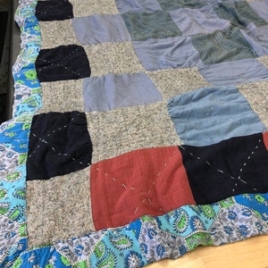 May include: A patchwork quilt featuring squares of blue, grey, red, and black fabrics. The quilt has a paisley border in blue and green. The quilt is likely made of cotton or a similar material, and is handmade.