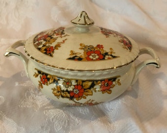 May include: A cream-colored ceramic sugar bowl with a matching lid, featuring two side handles and a gold-trimmed finial. The bowl and lid are decorated with a floral pattern in shades of orange, white, and black, accented with gold leaf details.