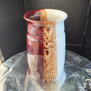May include: A cylindrical ceramic vase featuring a split-color design with a glossy deep red finish on one side and a smooth white finish on the other. A vertical band of textured, mottled brown glaze runs down the center, separating the two colors.