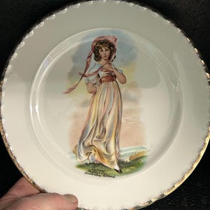 May include: A white ceramic plate with a gold rim features a painted image of a woman in a pink hat and a yellow dress. The plate is decorated with a gold rim and has the text "Kavel 107 Muller & Zoon Amsterdam Holland" printed on the bottom.