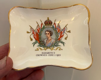 Puede incluir: Un plato rectangular de cerámica blanquecina con borde dorado. Presenta un retrato de la reina Isabel II, una corona y el texto "ELIZABETH II CROWNED JUNE 2 1953". El diseño incluye la bandera británica.