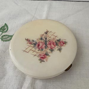 May include: A round, white compact mirror with a floral design. The design features pink roses and green leaves on a white background with a grid pattern.