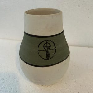 May include: A small, handcrafted ceramic vase. It has a cream-coloured top and bottom, and a sage green band in the middle. The green band features a black graphic of a feather within a circle. The vase has a rounded body and a cylindrical opening.
