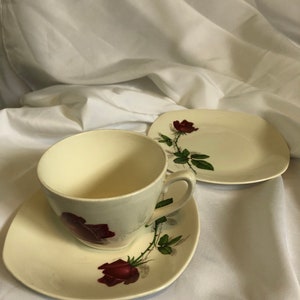 Könnte beinhalten: Ein cremefarbenes Tee-Set mit Tasse, Untertasse und kleinem Teller. Jedes Teil hat ein Design mit roten Rosen und grünen Blättern und Stielen. Die Tasse hat einen Henkel, und die Untertasse und der Teller haben eine geschwungene Form.