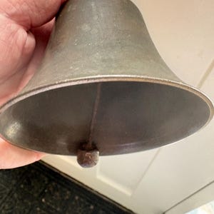 May include: A vintage metal bell with a wooden handle. The bell has a tarnished, aged appearance, with a rounded base and a clapper visible inside. The handle is a dark brown color.
