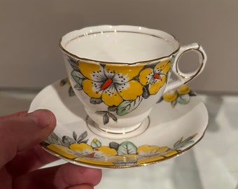 May include: A white porcelain teacup and saucer set featuring a vibrant yellow floral pattern with grey leaves and orange accents. The rim of both the cup and the saucer is finished with a thin gold trim, and the cup includes a small, curved handle.