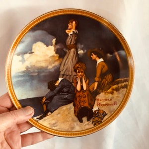 May include: Decorative plate featuring a painting by Norman Rockwell. The artwork depicts four figures in vintage clothing, set against a stormy sky. The plate has a gold rim and is held by a hand.