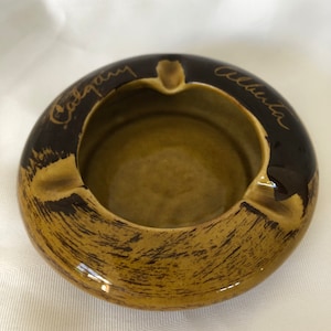 May include: A ceramic ashtray with a circular design. The ashtray has a brown exterior with a textured pattern and a lighter brown interior. The words "Calgary" and "Alberta" are written on the rim in a gold color.