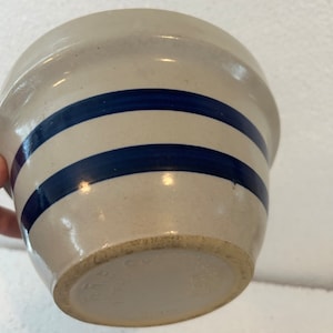 Puede incluir: Un cuenco de gres con base beige claro y dos rayas horizontales azul oscuro. El cuenco tiene una forma ligeramente acampanada y un acabado liso y brillante. La parte inferior del cuenco tiene texto estampado.