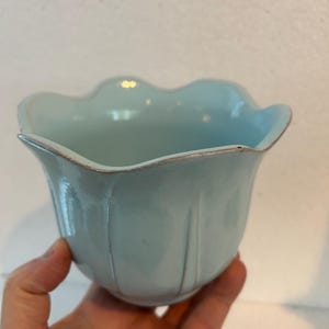 May include: A light blue ceramic bowl with a scalloped rim. The bowl has vertical ridges and a slightly textured surface. The rim has a subtle brown accent. The bowl is held in a hand.