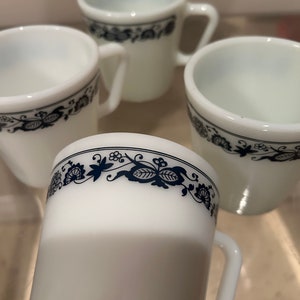 Puede incluir: Cuatro tazas de cerámica blanca con diseños florales azules alrededor de la parte superior. Las tazas tienen una forma redondeada y un asa.
