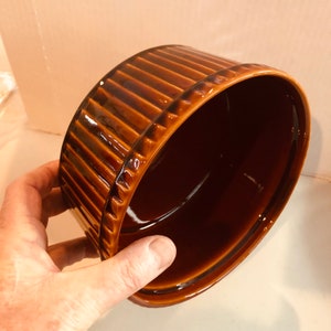May include: A brown ceramic oval baking dish with a ribbed exterior. The dish has a glossy finish and is held at an angle, showcasing its interior. The color is a rich, warm brown, suggesting a vintage or rustic aesthetic.
