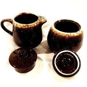 Pode incluir: Um conjunto vintage de cremeira e açucareiro de cerâmica marrom com tampas. A cremeira tem uma alça e um bico. O açucareiro é redondo. Ambas as peças têm um esmalte marrom com uma borda marrom mais clara.