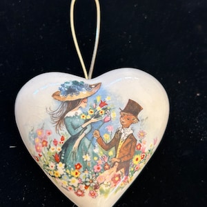 May include: Heart-shaped ceramic ornament with a painted scene of a couple in a flower garden. The ornament has a gold-colored wire loop for hanging. The artwork features a woman in a blue dress and hat and a man in a brown suit.