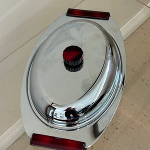 May include: A silver oval serving dish with a red knob on the lid. The dish has two red handles on the sides.