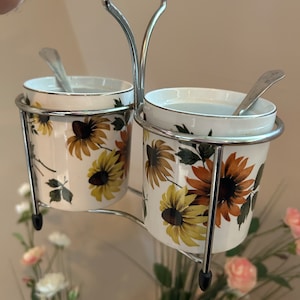 Può includere: Un portacondimenti doppio vintage con due piccoli contenitori in ceramica. Ogni contenitore presenta un motivo floreale di girasole in giallo, marrone e verde su sfondo bianco. I contenitori hanno bordi argentati e cucchiaini, tenuti in un supporto in metallo argentato.
