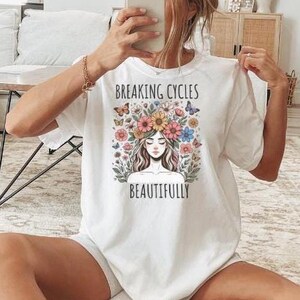 May include: White t-shirt with the text "BREAKING CYCLES BEAUTIFULLY" above a floral illustration of a woman's face. The shirt is worn with white socks with black stripes. The person is sitting on a rug.
