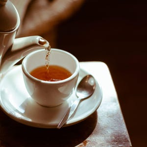 K&ouml;nnte beinhalten: Eine wei&szlig;e Teekanne gie&szlig;t Tee in eine wei&szlig;e Teetasse mit Untertasse. Die Teetasse steht auf einem Holztisch.