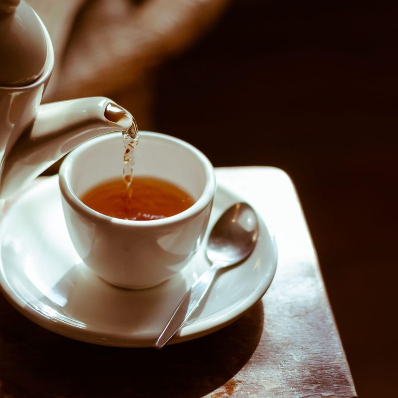 K&ouml;nnte beinhalten: Eine wei&szlig;e Teekanne gie&szlig;t Tee in eine wei&szlig;e Teetasse mit Untertasse. Die Teetasse steht auf einem Holztisch.