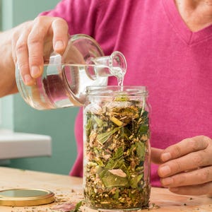 May include: A person pouring clear liquid from a glass bottle into a glass jar filled with dried herbs and flowers.