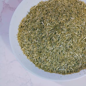 May include: A white plate filled with dried, chopped green herbs. The herbs have a light green and tan color, with a texture of small, thin pieces. The plate is set against a white and gray marble surface.