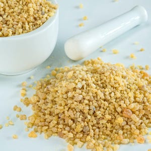 May include: A white ceramic mortar and pestle next to a pile of golden-yellow granules. A matching bowl is filled with the same granules. The granules have a slightly translucent appearance and vary in size. The background is a light blue.