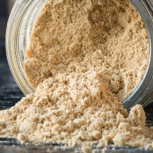 May include: A glass jar filled with a light brown powder, spilling out onto a dark surface. The powder is fine and granular, with a slightly rough texture.