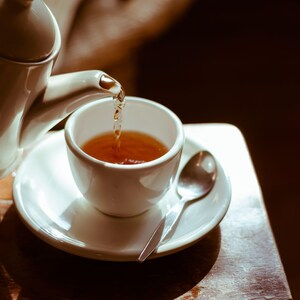 May include: A white teapot pouring tea into a white teacup with a saucer. The teacup is filled with brown tea. A silver spoon is on the saucer.