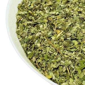 May include: A close-up of a white bowl filled with dried, chopped, green moringa leaves.