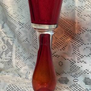 May include: A tall, ruby red glass candlestick holder featuring a flared top, a slender neck with a bulbous center, and a wide, circular base. The glass transitions from a deep, saturated red at the top and bottom to a slightly lighter hue.