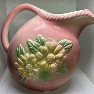 Puede incluir: Una jarra de cerámica rosa con un diseño floral tridimensional en relieve con pétalos y hojas amarillas y verdes. La jarra tiene un asa curva, una textura similar a una cuerda a lo largo del borde, un acabado brillante y un cuerpo redondeado.