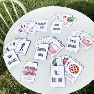 May include: A selection of educational flashcards with apple-themed illustrations and text. The cards are arranged on a white table and include categories like vocabulary, sequencing, and spatial concepts. The cards feature images of apples, baskets, and related items.
