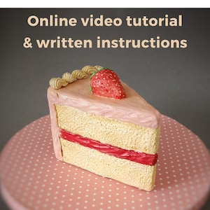 May include: A slice of cake with a strawberry on top, displayed on a pink polka dot surface. The cake has a light pink frosting and a red filling. Text at the top reads: Online video tutorial & written instructions.