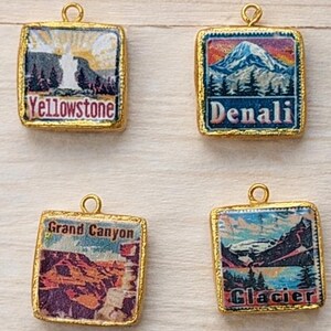 May include: A collection of nine square gold-rimmed charms, each featuring a colorful illustration of a U.S. National Park. The charms are labeled with the names Rocky Mountain, Yellowstone, Denali, White Sands, Grand Canyon, Glacier, Joshua Tree, Sequoia, and Haleakala.