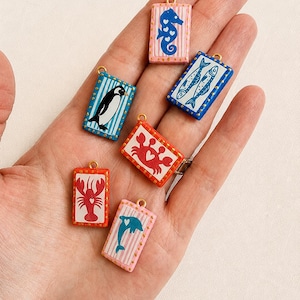 May include: A collection of rectangular charms featuring marine life illustrations. The charms include a seahorse, fish, penguin, crab, lobster, and dolphin. Each charm has a gold-colored loop and is painted with bright colors and striped backgrounds.